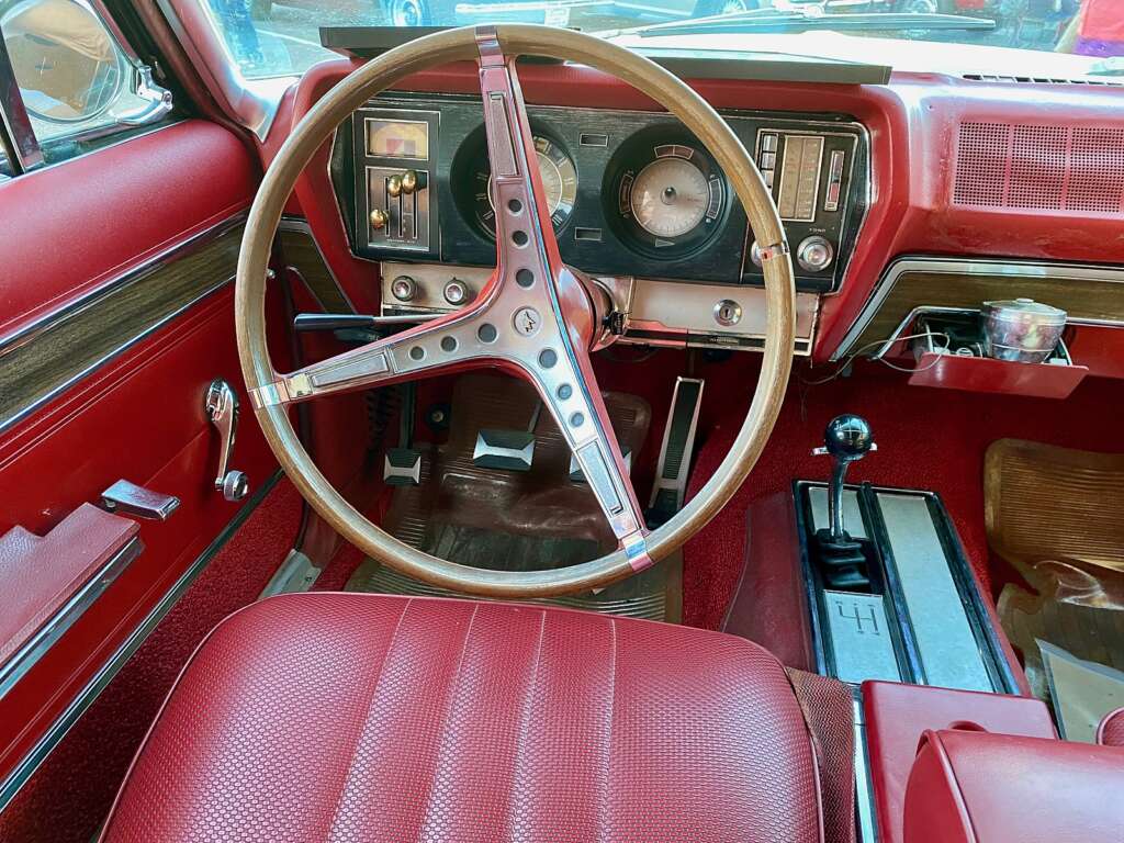 A vintage car interior featuring a red leather bench seat, a wooden steering wheel, and a classic dashboard with analog gauges. The manual gear shift is situated on the floor next to a console, surrounded by red carpet and trim.
