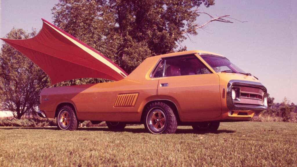 A vintage car with an unusual design features a large, wing-like structure extending from its rear. The car is parked on a grassy area with trees in the background, under a clear sky. The vehicle appears to be from the mid-20th century, painted in an orange hue.