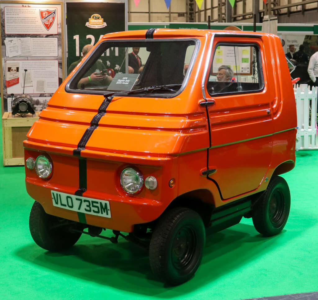 An orange, three-wheeled microcar is displayed in an indoor exhibition with information boards and equipment in the background. The vehicle has a compact design, round headlights, and a front license plate reading "VLO 735M." The floor is covered in green carpet.