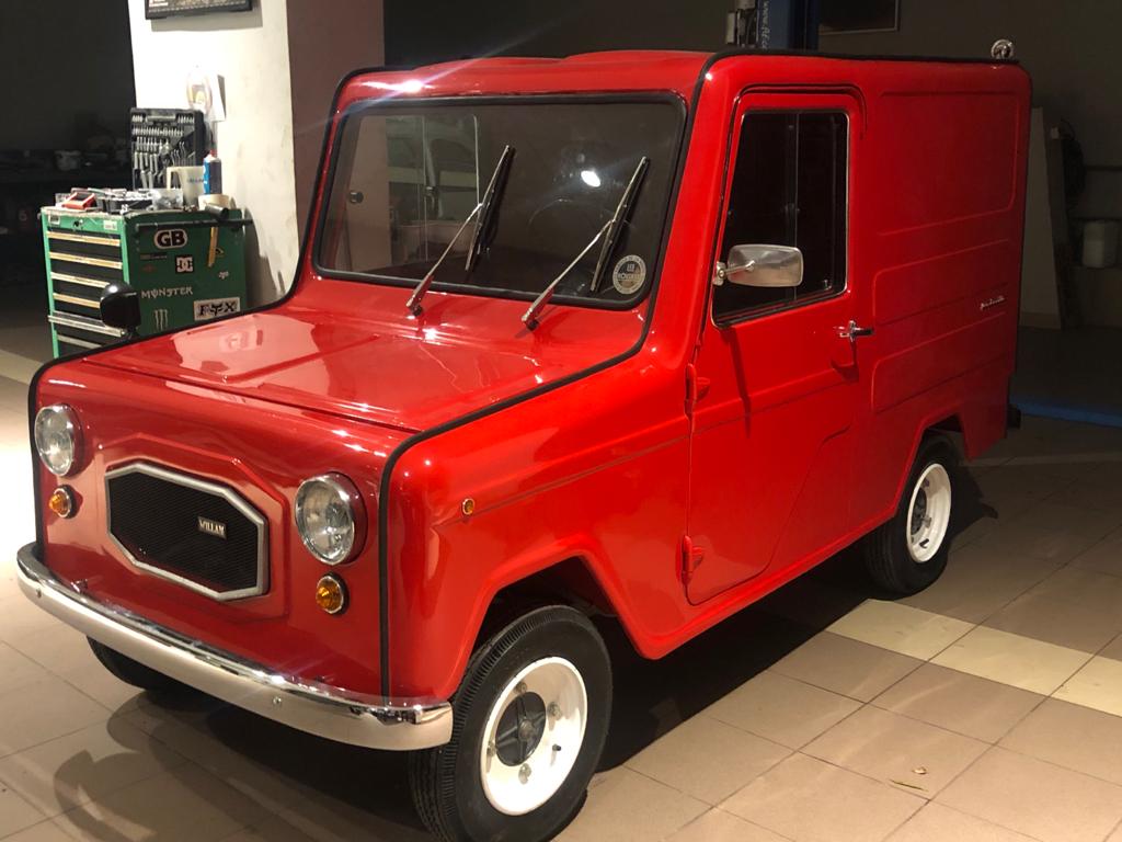 A small, vintage red van with a boxy shape is parked indoors on a tiled floor. Its design features a prominent grille with the brand name "VISANI" and round headlights. The van has white-rimmed wheels and is set against a backdrop of a workshop or garage.