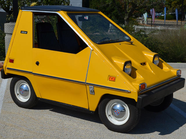 A small, yellow, wedge-shaped car with a compact body and two round headlights is parked outdoors. The car features a flat roof, narrow tires, and a minimalistic design, with its brand name "CitiCar" visible on the side. Trees and a fence are in the background.