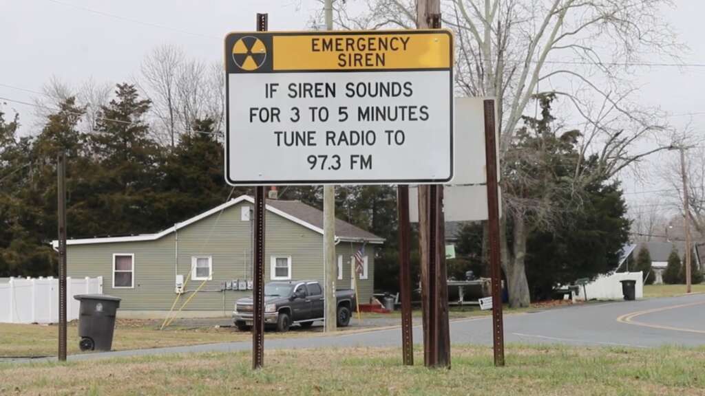 A street sign in a suburban area reads, "EMERGENCY SIREN. IF SIREN SOUNDS FOR 3 TO 5 MINUTES TUNE RADIO TO 97.3 FM." The sign is mounted on metal poles next to a street, with houses and trees visible in the background.