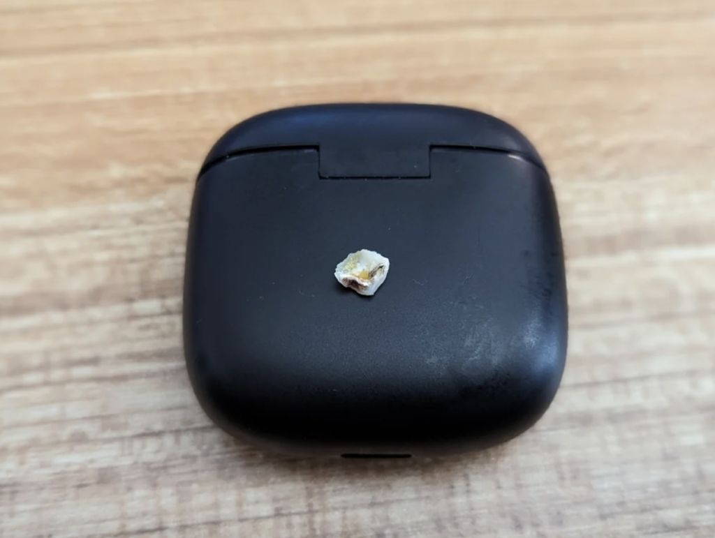 A small gold and white stone rests on a closed black earbuds case, placed on a wooden surface.
