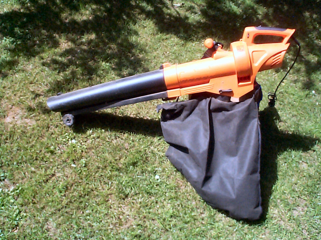 An orange leaf blower vacuum sits on a grassy surface. It has a extended black nozzle and a large black collection bag attached to its side. Sunlight casts shadows from nearby trees.