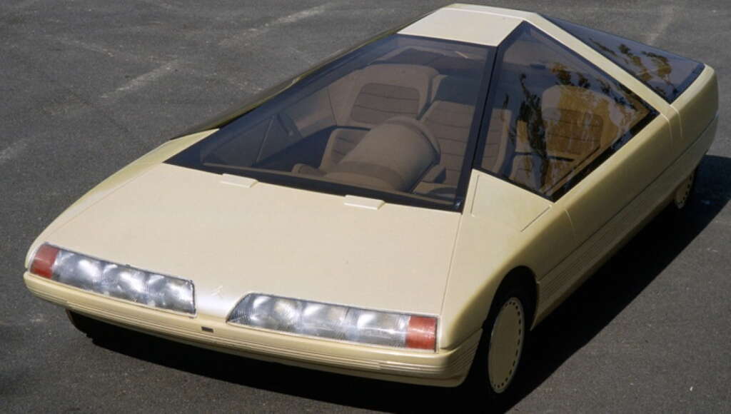 A futuristic beige concept car with a sharply angular, low-profile design, featuring a large, sloped windshield that extends over the roof, and distinctive rectangular headlights. The car has a sleek, minimalist appearance, set against a plain background.