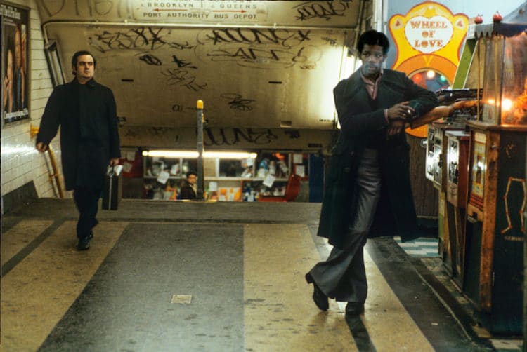 In a dimly lit subway station covered in graffiti, a man in dark clothing walks towards another man who is leaning against a "Wheel of Love" machine. The scene has a gritty, urban atmosphere with other people visible further back in the tunnel.