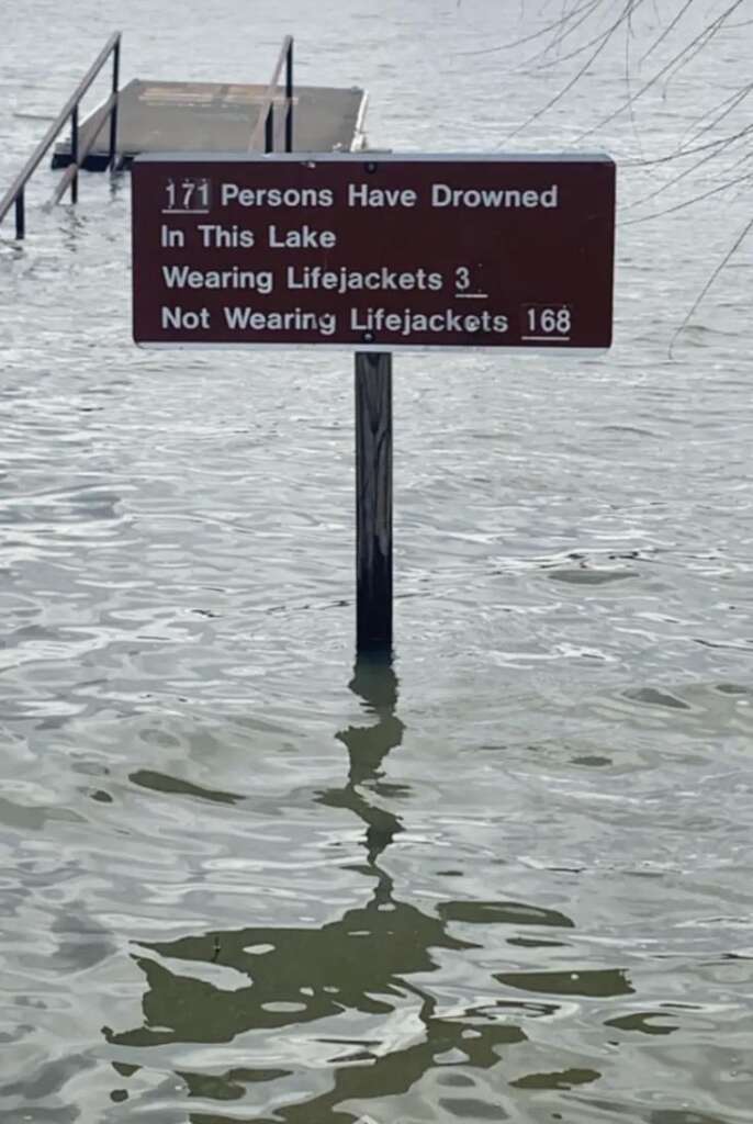 A partially submerged sign in a lake reads: "171 Persons Have Drowned In This Lake. Wearing Lifejackets: 3. Not Wearing Lifejackets: 168." A pier or dock can be seen extending from the water in the background. The water level is high.