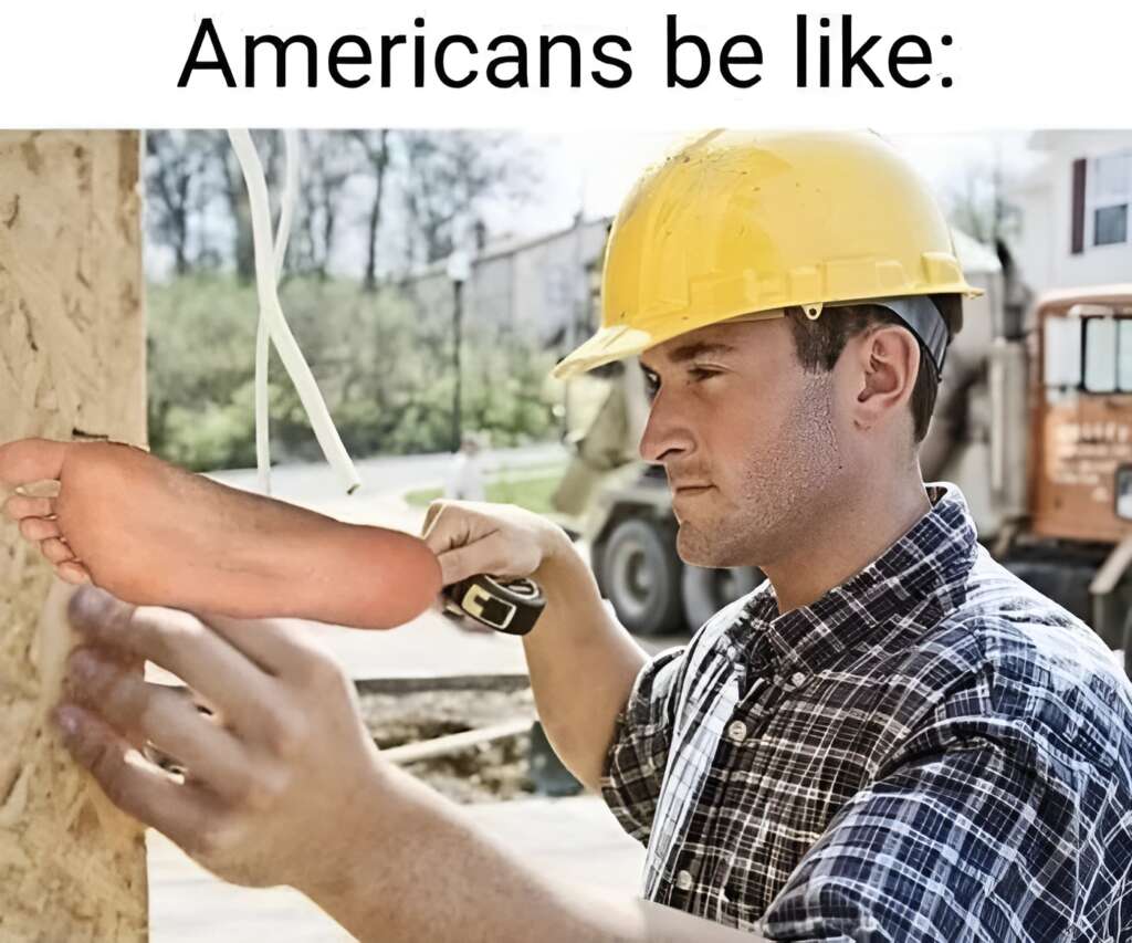 A construction worker wearing a hard hat is hammering a large foot onto a wooden surface. The text at the top reads, "Americans be like: