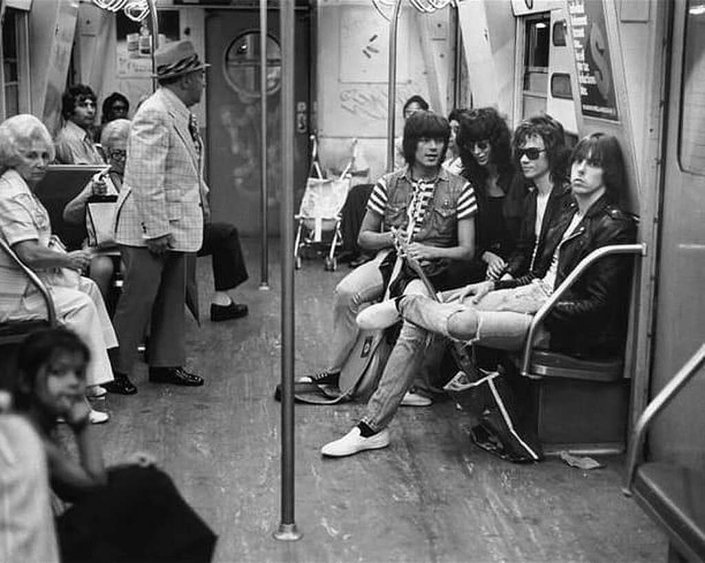 A black and white photo of a subway car. A group of four individuals with long hair and wearing sunglasses, casual clothes, and leather jackets sit together on the right. Other passengers include older individuals seated on the left and a man standing near the door.