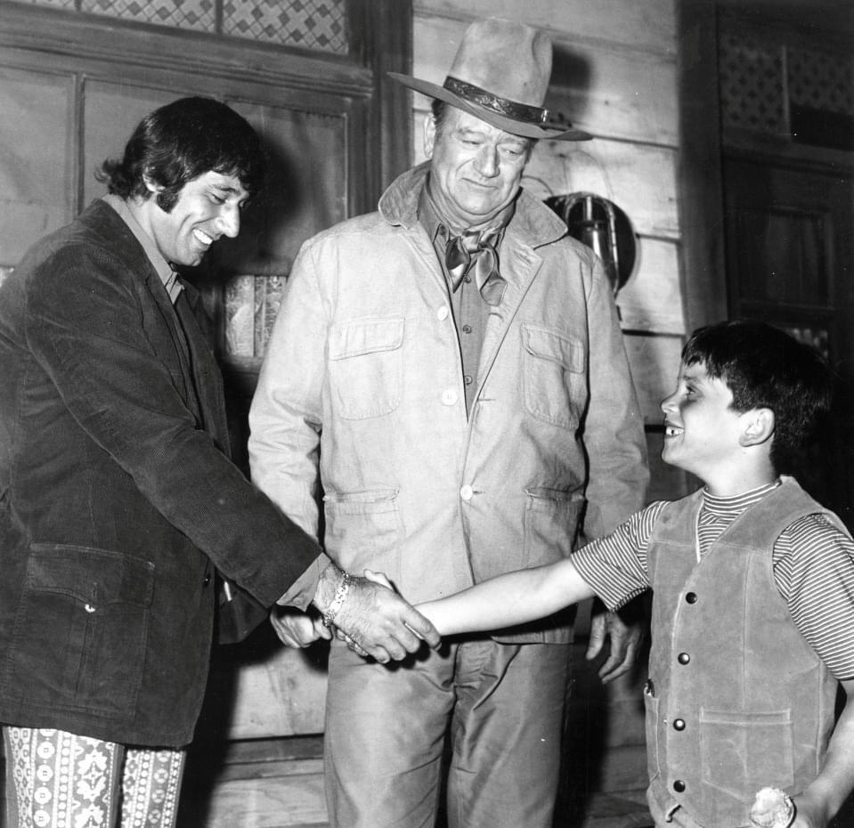 A man in patterned pants shakes hands with a boy in a striped shirt and vest as an older man in a cowboy hat and jacket looks on smiling. The three of them are standing in front of a rustic wooden building.