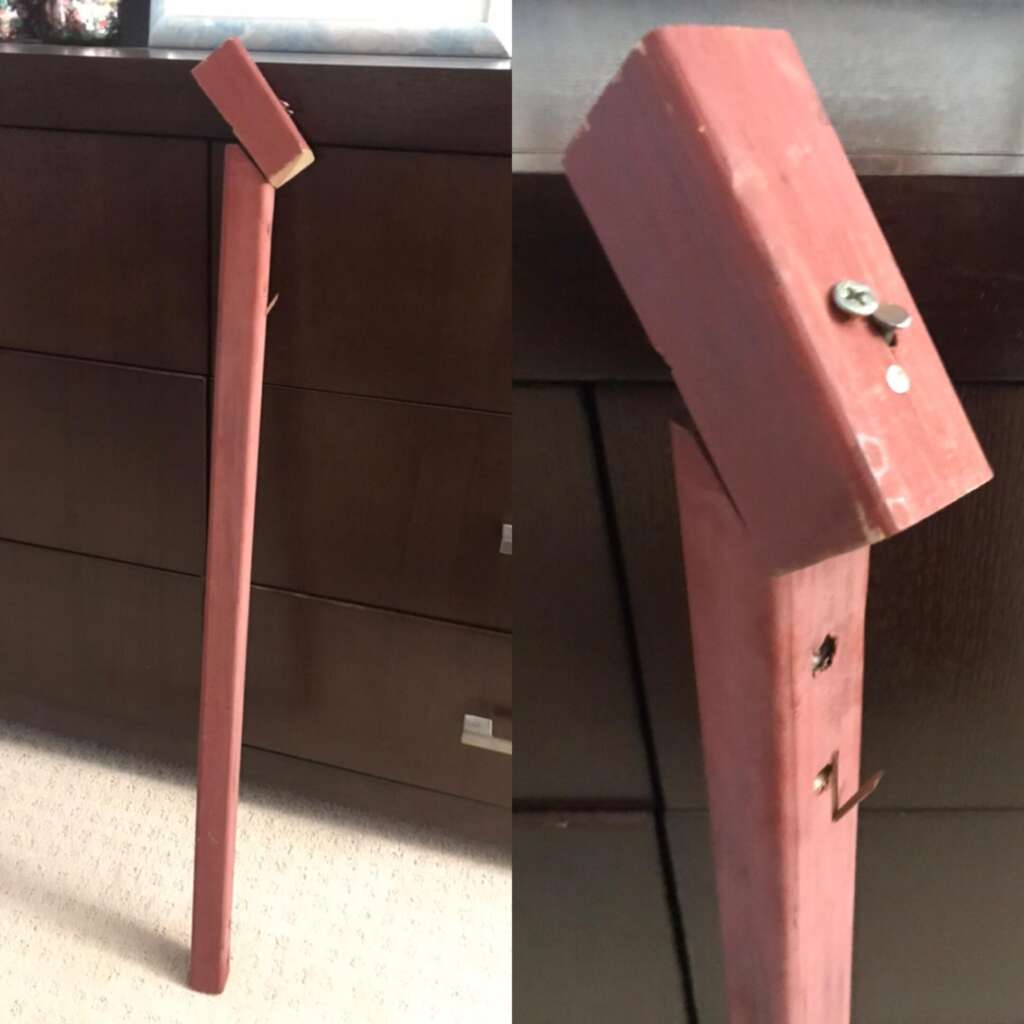 A wooden stick painted red, leaning against a dark brown dresser. The stick has a diagonal top end with a screw and a hook attached to it. In the close-up view, the screw and hook details are more visible. The stick appears to be handmade and weathered.