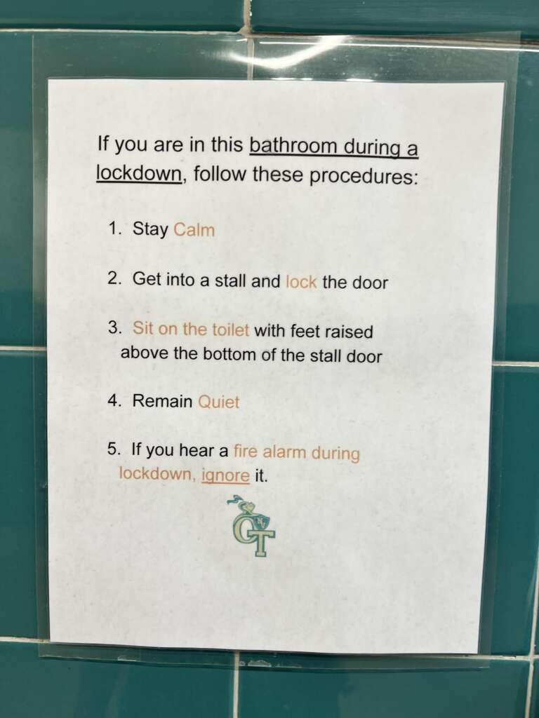 A printed sign on a teal-tiled wall outlines bathroom lockdown procedures. It states to stay calm, get into and lock a stall, sit on the toilet with feet raised, remain quiet, and ignore fire alarms during the lockdown. The logo "CT" is at the bottom.