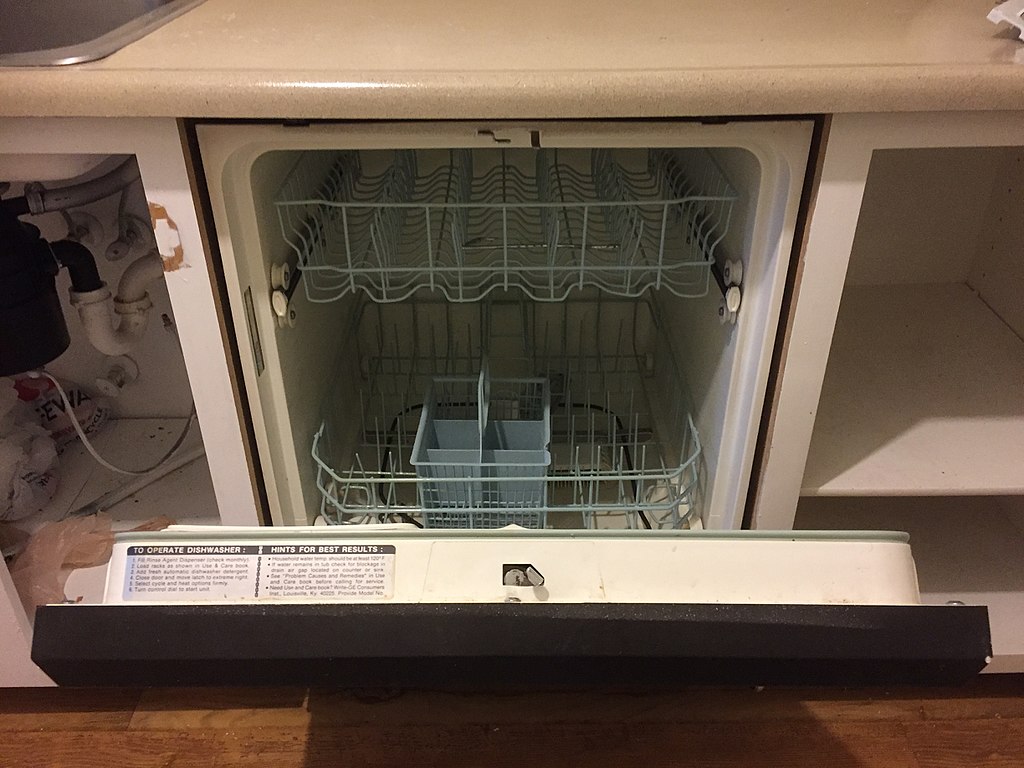 Open dishwasher with the door fully lowered. The interior is empty and features two racks and a silverware basket. The surrounding kitchen cabinetry is white, and the countertop is beige. Some plumbing is visible under the sink to the left.