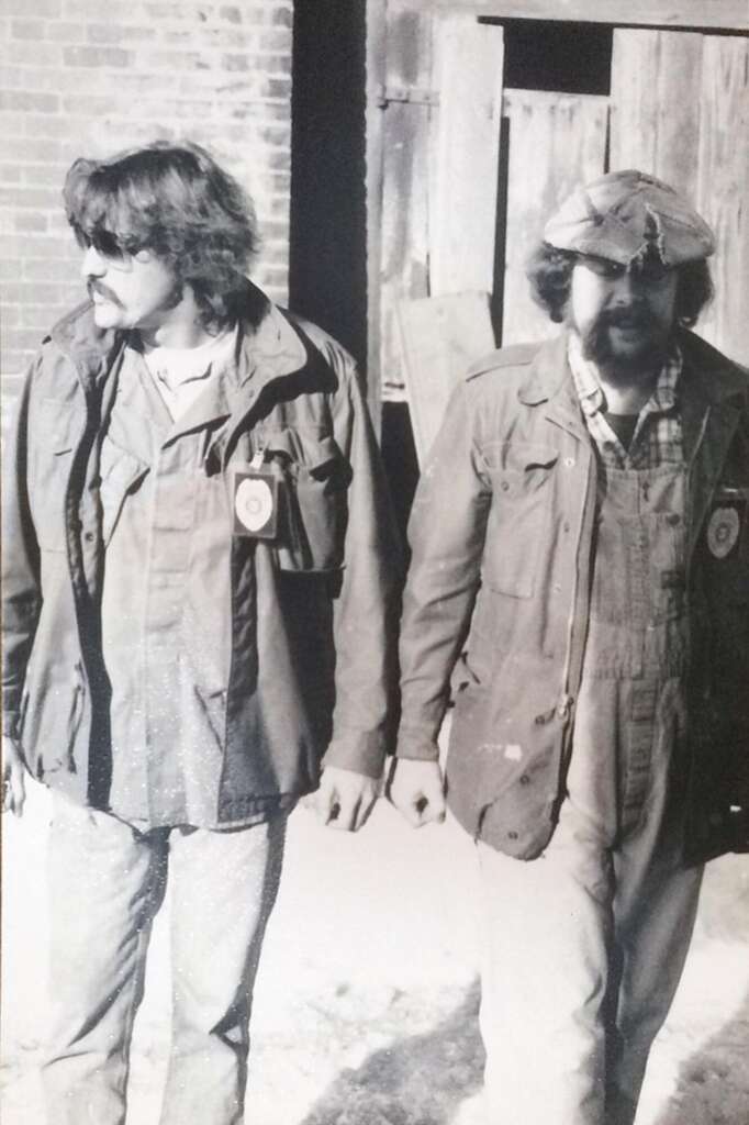 Two men with beards and long hair walk side by side in an outdoor setting, both dressed in casual layered clothing, including jackets and hats. The background shows a brick wall and a wooden door. The image is in black and white, giving it a vintage feel.