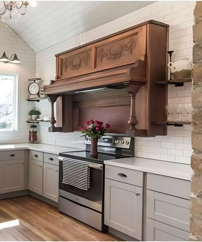 A farmhouse-style kitchen features an ornate, wooden range hood above a stainless steel oven. Cabinets and drawers are painted white and grey. The back wall is adorned with white subway tiles. A vase with red flowers sits on the counter next to the oven.