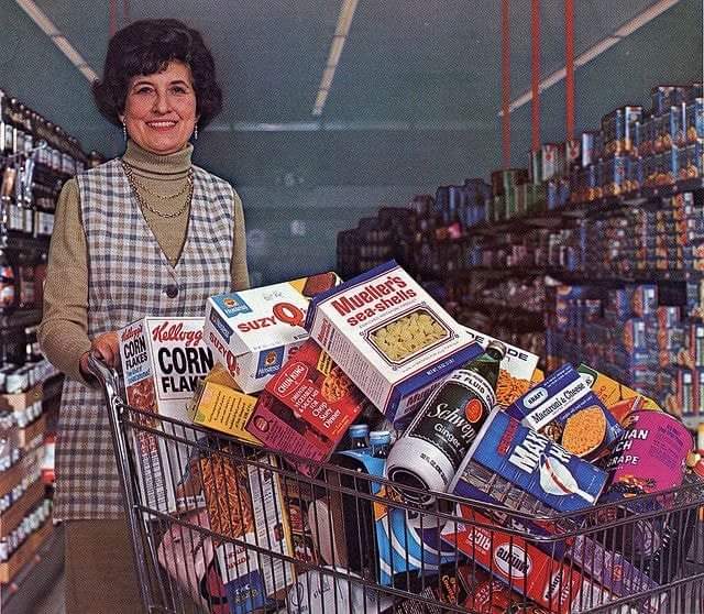 A woman with short curly hair and a checkered dress smiles while pushing a shopping cart filled with groceries. The cart contains items like cereal boxes, crackers, and canned goods. She stands in a brightly lit grocery store aisle lined with shelves.