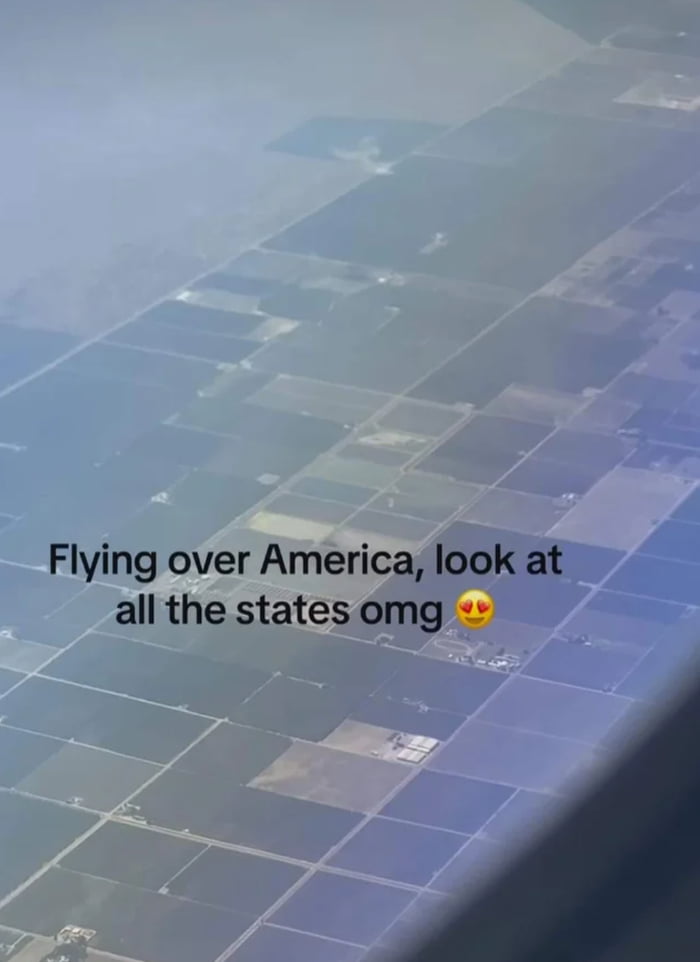 View of a patchwork landscape from an airplane window, showing farmland and fields below. Overlaid text reads, "Flying over America, look at all the states omg" followed by a heart-eyed emoji. The photo captures a mix of land and some cloud cover.