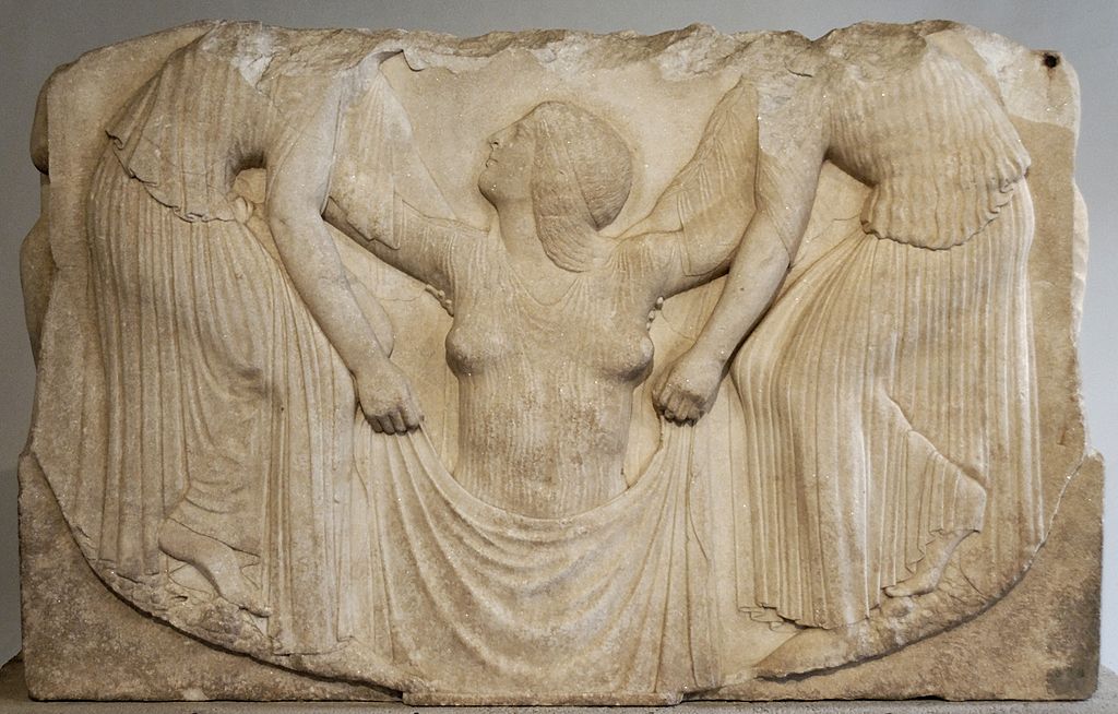 A classical marble relief sculpture depicts three draped figures. The central figure appears to be either emerging from or being wrapped in fabric held by the two other figures, each standing on either side, displaying detailed folds in their garments.