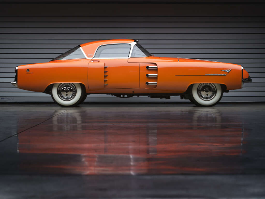 A sleek orange vintage car with a streamlined design is parked indoors against a gray horizontal paneled wall. The car has white wall tires and a distinctive curved roof. Elegant reflections of the car can be seen on the shiny floor.