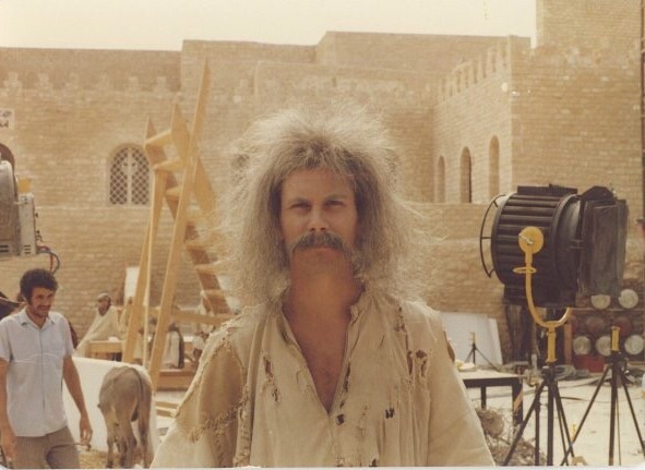 A man with wild, gray, frizzy hair and a thick mustache stands in front of a film set with stone buildings and large production lights. He is wearing a tattered beige outfit. In the background, another man and a donkey can be seen.