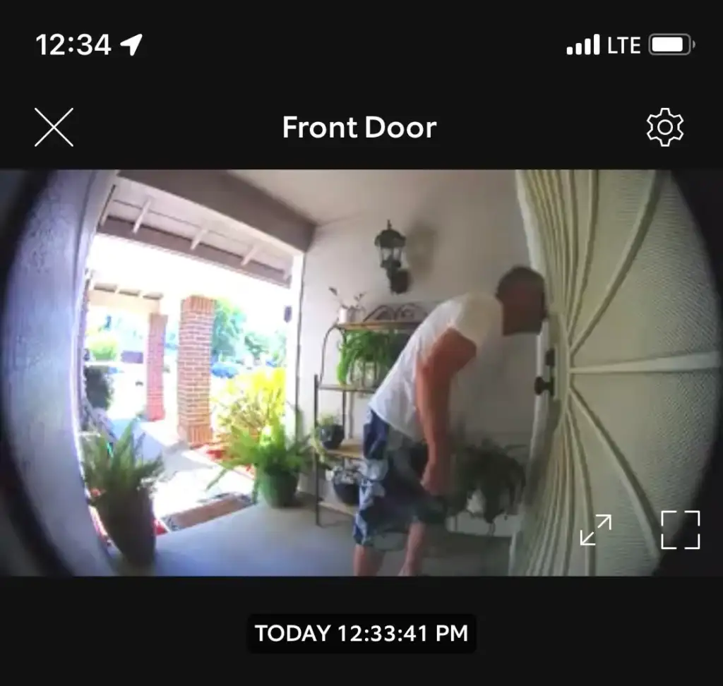 Image from a doorbell camera showing a man standing at the front door. He is examining the door, partially bent towards it, and is dressed in a white t-shirt and shorts. The time stamp on the image reads "TODAY 12:33:41 PM". Several plants adorn the porch.