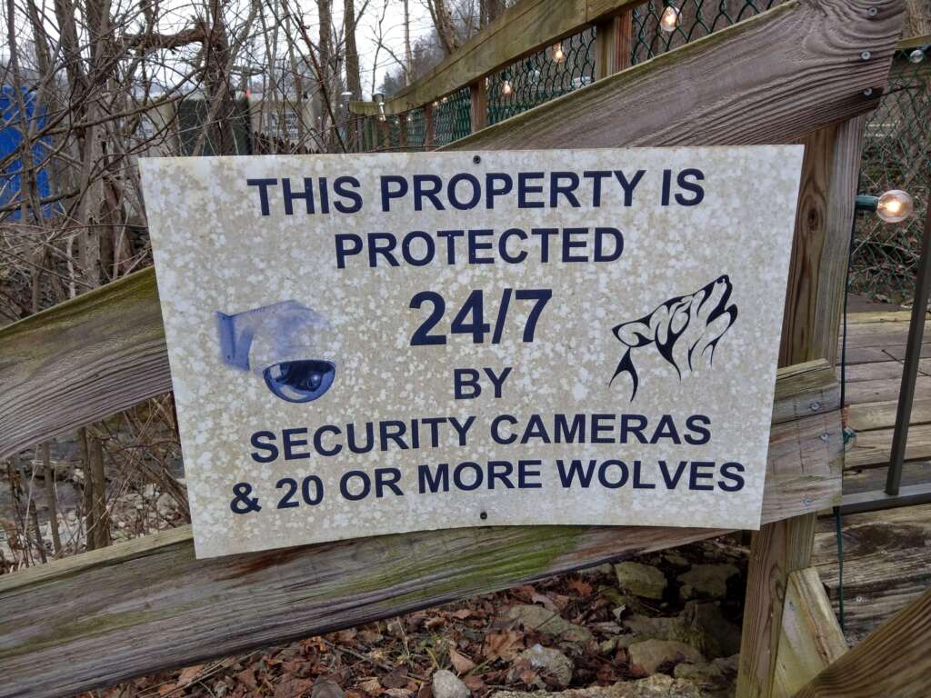 A sign posted on a wooden fence reads, "This property is protected 24/7 by security cameras & 20 or more wolves," with images of a surveillance camera and a wolf howling. The background shows trees and a wire fence.