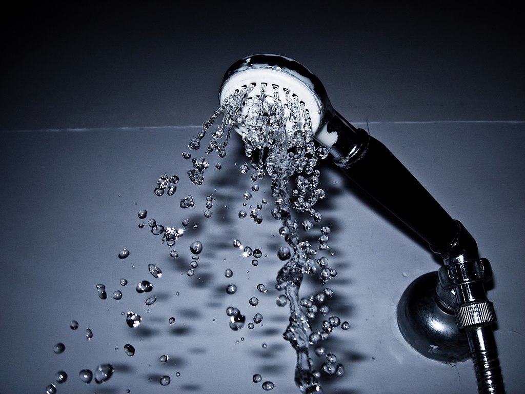 Close-up of a showerhead spraying water. The water droplets are captured mid-air against a dark background, highlighting their clarity and movement. The showerhead is silver with a black handle.