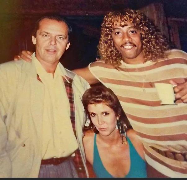 Three people are posing together in a casual setting. The person on the left is wearing a light-colored jacket. The person in the middle, a woman with shoulder-length hair, is wearing a blue top. The person on the right, with curly hair, is wearing a striped shirt.