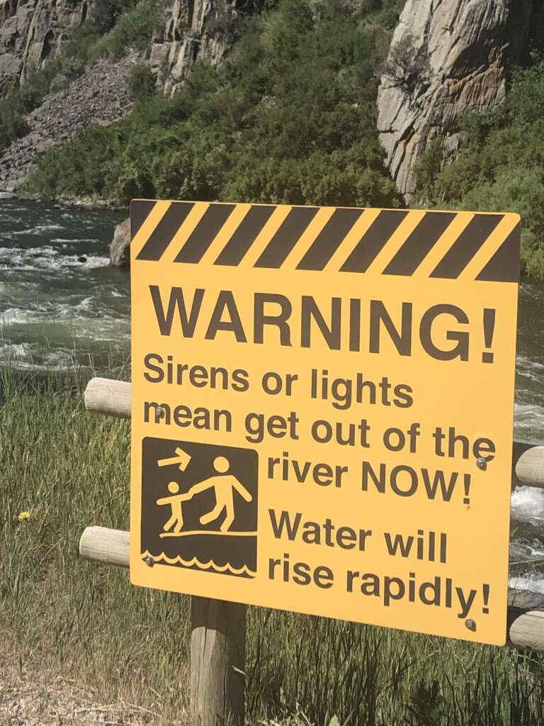 A yellow warning sign with black text and striped border is posted near a river. It reads, "WARNING! Sirens or lights mean get out of the river NOW! Water will rise rapidly" and includes a pictogram of a person hurriedly exiting water. Rocky terrain and vegetation are visible in the background.