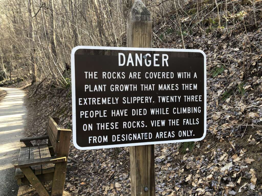 A warning sign on a wooded trail reads, "DANGER: The rocks are covered with a plant growth that makes them extremely slippery. Twenty three people have died while climbing on these rocks. View the falls from designated areas only." A bench is nearby.