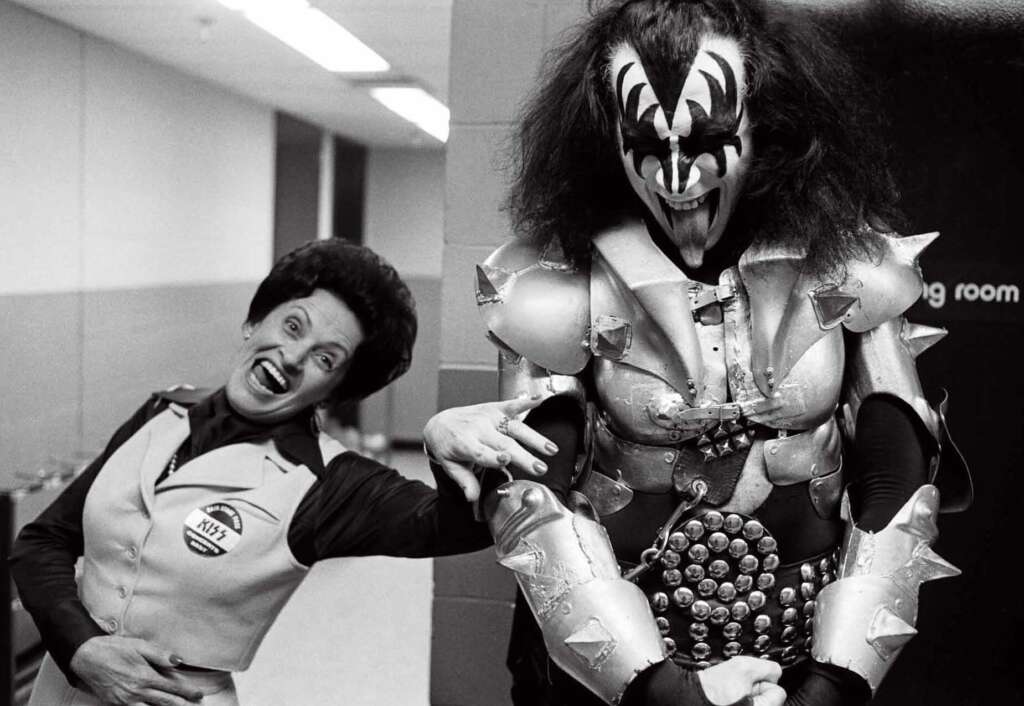 A woman excitedly poses next to a man dressed in an elaborate rock costume, complete with face paint, spikes, and leather. The pair stand in a corridor, with the man appearing in character with an outstretched tongue and dramatic facial expression.