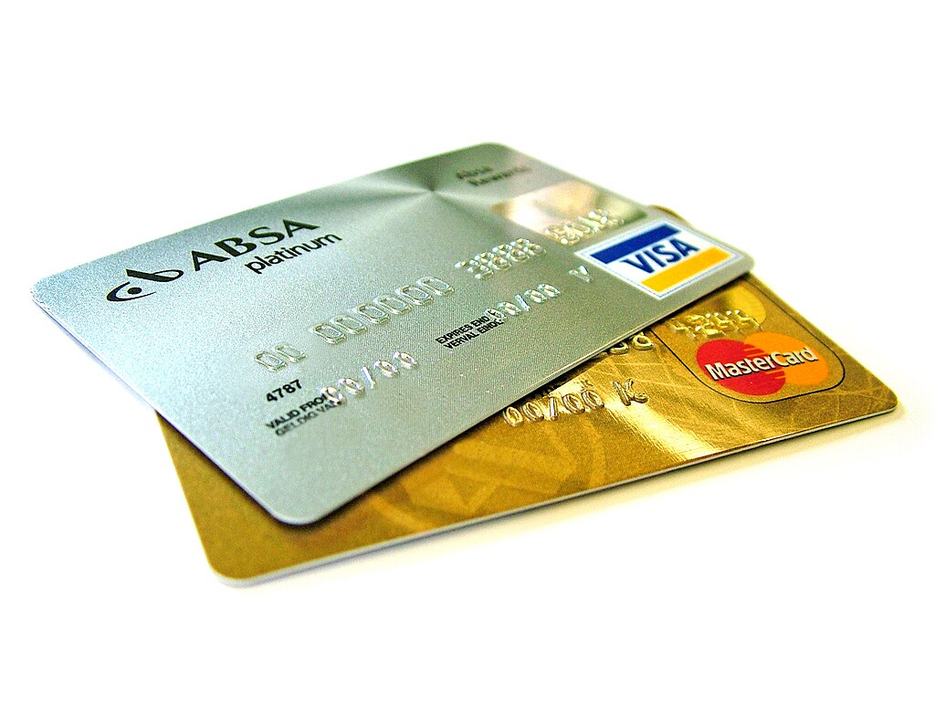 Close-up of two credit cards layered on top of each other. The top card is a silver ABSA Platinum Visa card, and the bottom card is a gold MasterCard. Both cards have visible numbers, logos, and some text, though the details are out of focus. The background is white.