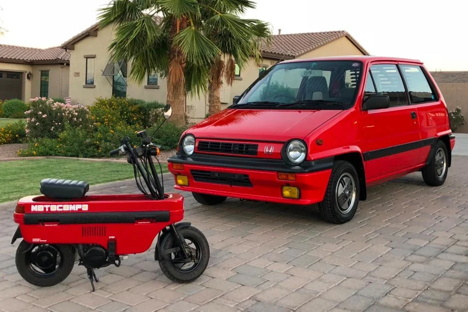 A red Honda City car is parked on a brick driveway in front of a house with palm trees. Next to the car is a red Honda Motocompo foldable scooter. Both vehicles are vintage and share a matching color scheme.