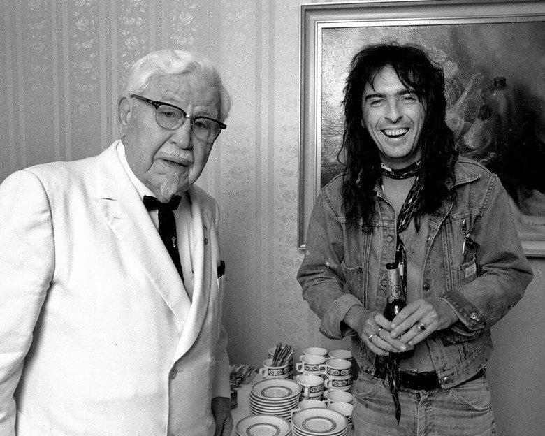 A black-and-white photo of an elderly man in a white suit, white shirt, black tie, and glasses, standing next to a younger man with long hair, wearing a denim jacket and accessorized with necklaces and bracelets, smiling and standing near a table with plates and cups.