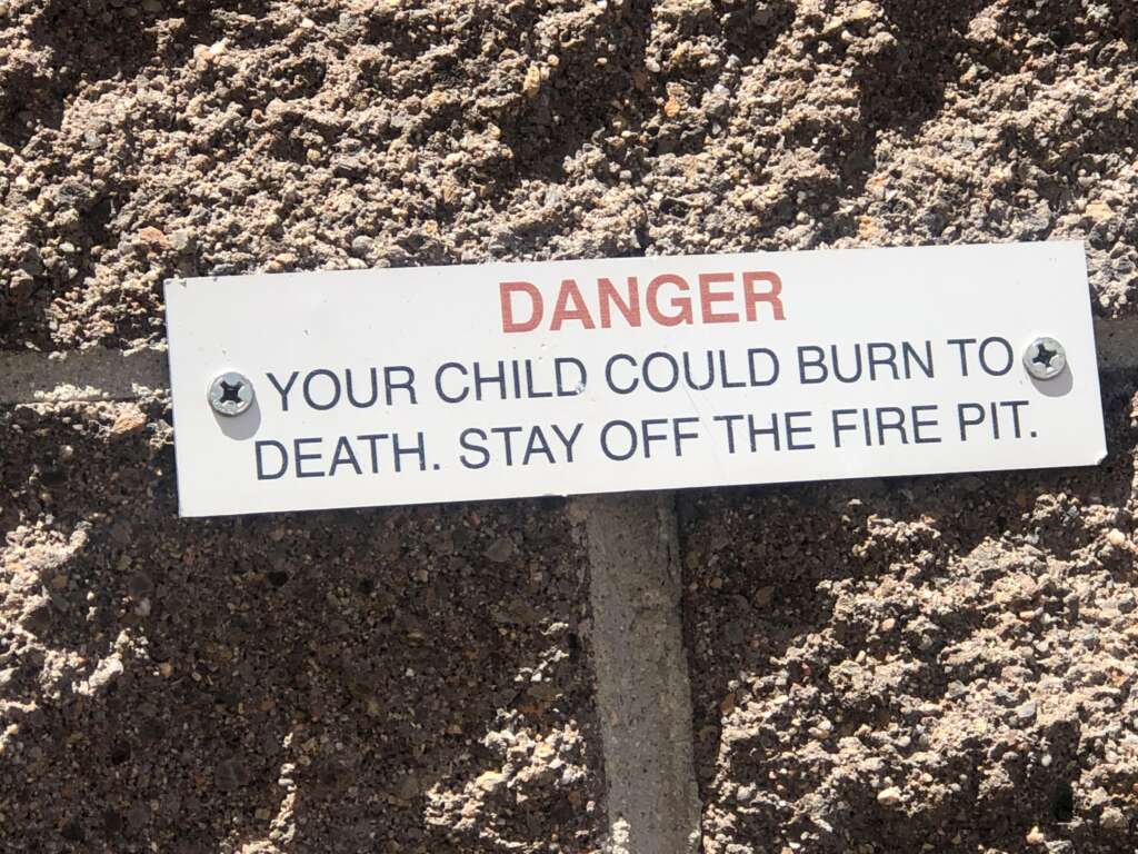 A warning sign is affixed to a rough, stone wall. The sign reads: "DANGER YOUR CHILD COULD BURN TO DEATH. STAY OFF THE FIRE PIT." The text is in black and red, with "DANGER" highlighted in red. The sign is secured with two screws.
