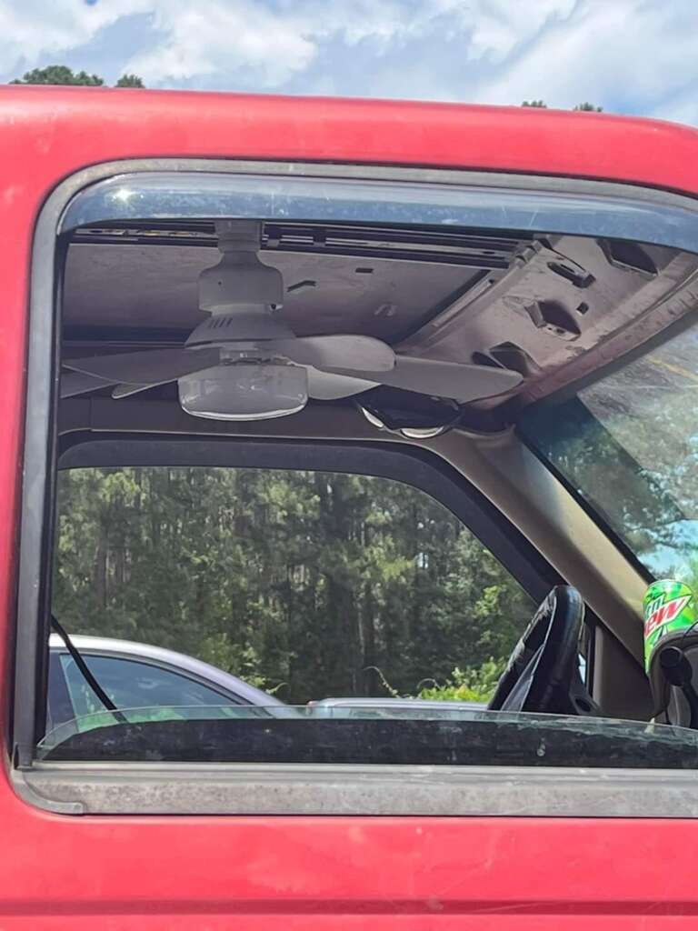 Close-up of a red vehicle's driver-side door with the window open. Inside, a white ceiling fan is installed on the underside of the vehicle's roof. A can of Mountain Dew is in the cup holder on the center console.