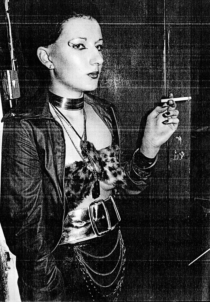 A person with dramatic eye makeup and short hair is standing against a dark wall. They are wearing a choker, layered necklaces, and a furry top, and are holding a cigarette in their right hand. Their attire includes a belt with chains and a dark jacket.