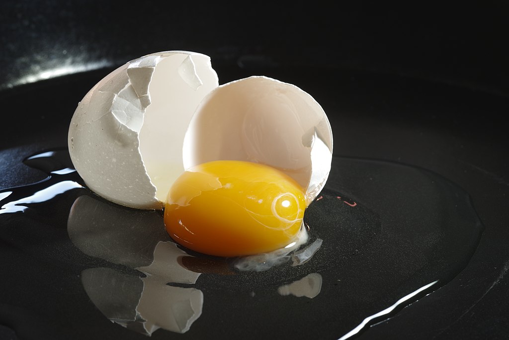 Close-up of a broken egg with the white shell and golden yolk spread out on a dark, smooth surface. The yolk is intact and shiny, surrounded by a translucent egg white. The overall background is dark, highlighting the egg's bright colors.