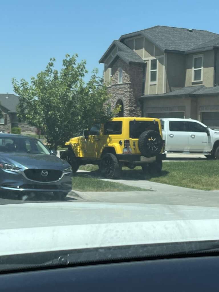 A yellow Jeep with a spare tire on the back is parked on the grass next to the driveway of a suburban house. A white truck is parked in the driveway, and a gray car is in front of the house. The sky is clear and blue.