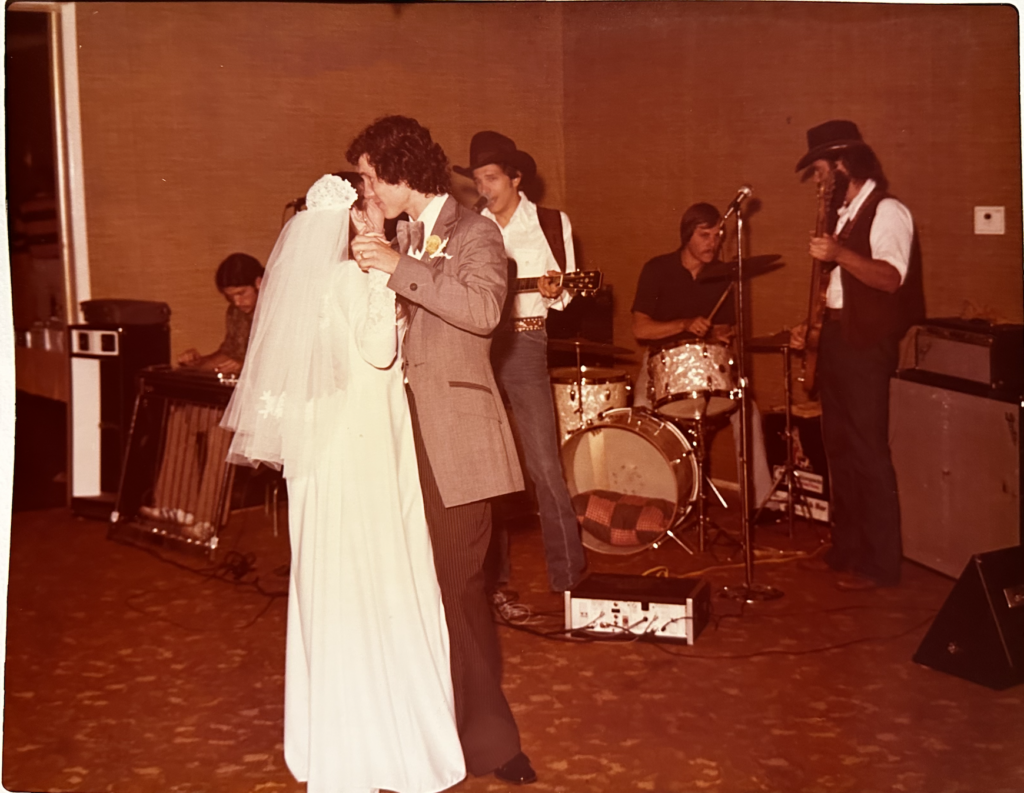 A bride and groom share their first dance at a wedding. The bride wears a white dress and veil, while the groom is in a gray suit. A live band with five members, including a drummer and guitarist, performs in the background. The venue has carpeted floors and beige walls.