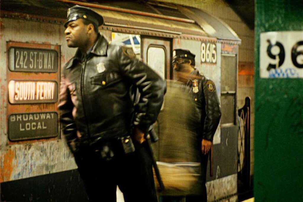 Two police officers in leather jackets and hats stand on a subway platform. One is in the foreground looking to the left, while the other stands in front of a stopped subway train, partially hidden. The train's sign reads "242 St - Broadway Local" and "South Ferry".