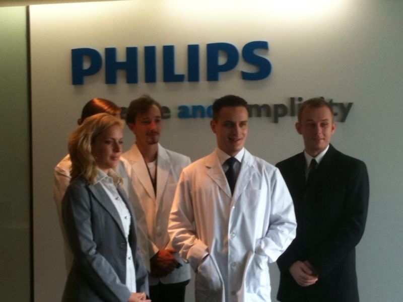 Four people stand in front of a wall with the Philips logo. Two individuals wear white lab coats, and the other two are dressed in formal business attire. They appear to be posing for a photo in a professional environment.