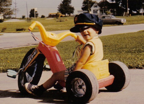 A young child wearing a large hat is seated on a tricycle in a suburban area. The child smiles at the camera, and there are houses, trees, and a parked car in the background. It appears to be a sunny day.