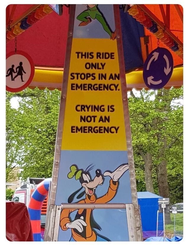 A colorful amusement ride sign features a cartoon character. The sign reads: "This ride only stops in an emergency. Crying is not an emergency." The cartoon character at the bottom appears to be waving with a cheerful expression.