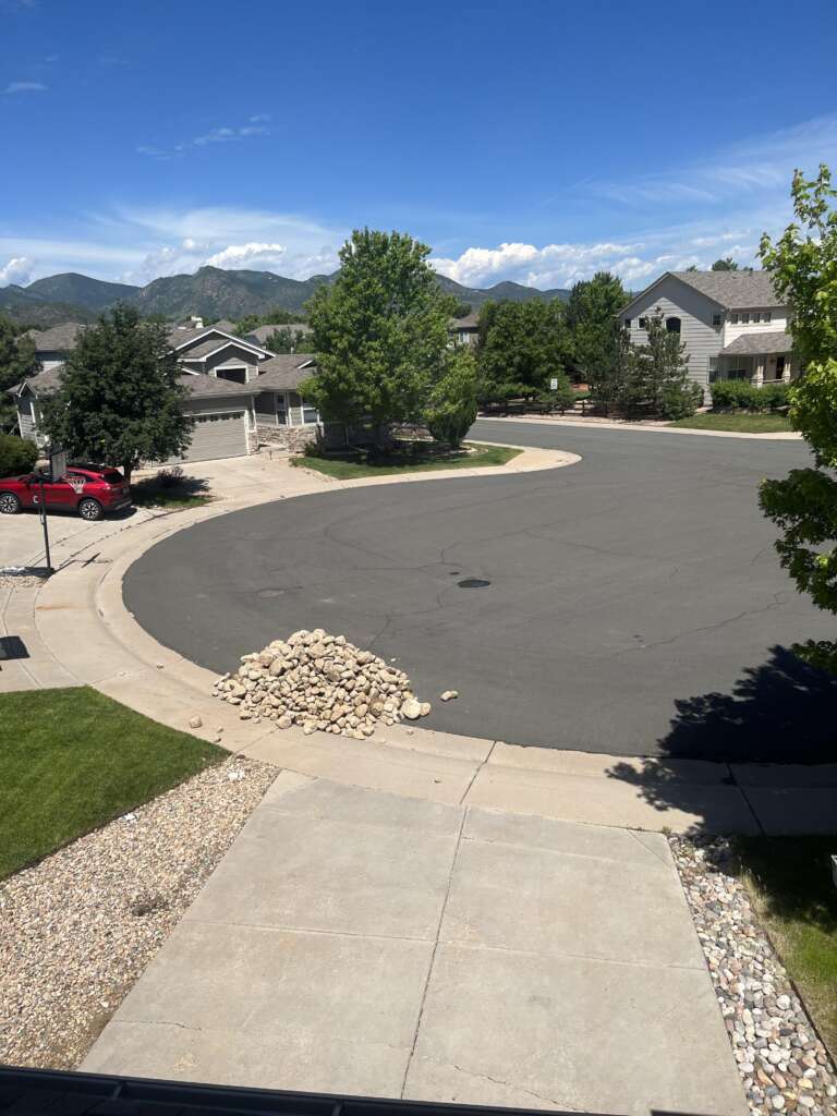 A suburban cul-de-sac with several houses surrounded by green trees and lawns. A pile of rocks is placed on the edge of the street, and a red car is parked in a driveway. Mountains are visible in the background under a clear blue sky.