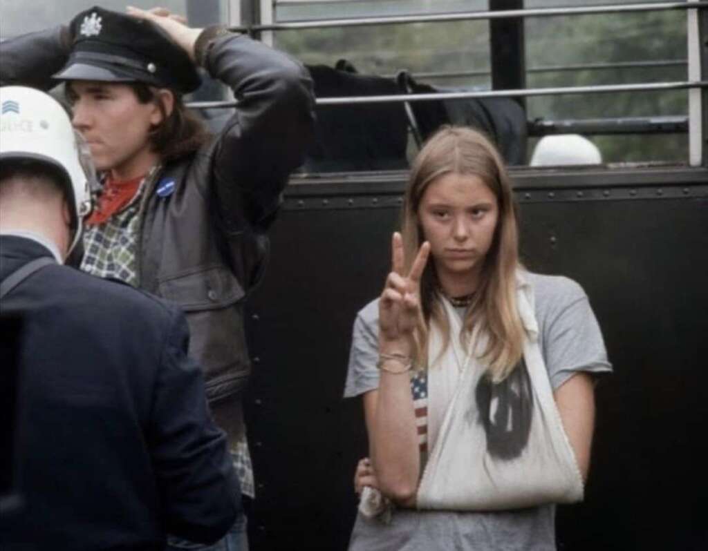 A young woman with a sling on her arm flashes a peace sign while standing next to a bus. To her right, a man in a leather jacket and hat is raising his right arm. In the foreground, a person in a police uniform is partially visible.