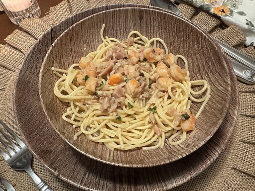 A wooden bowl filled with spaghetti topped with a seafood mix, including shrimp and pieces of fish, garnished with herbs. The bowl rests on a textured placemat, accompanied by a fork, knife, and a floral-patterned napkin.