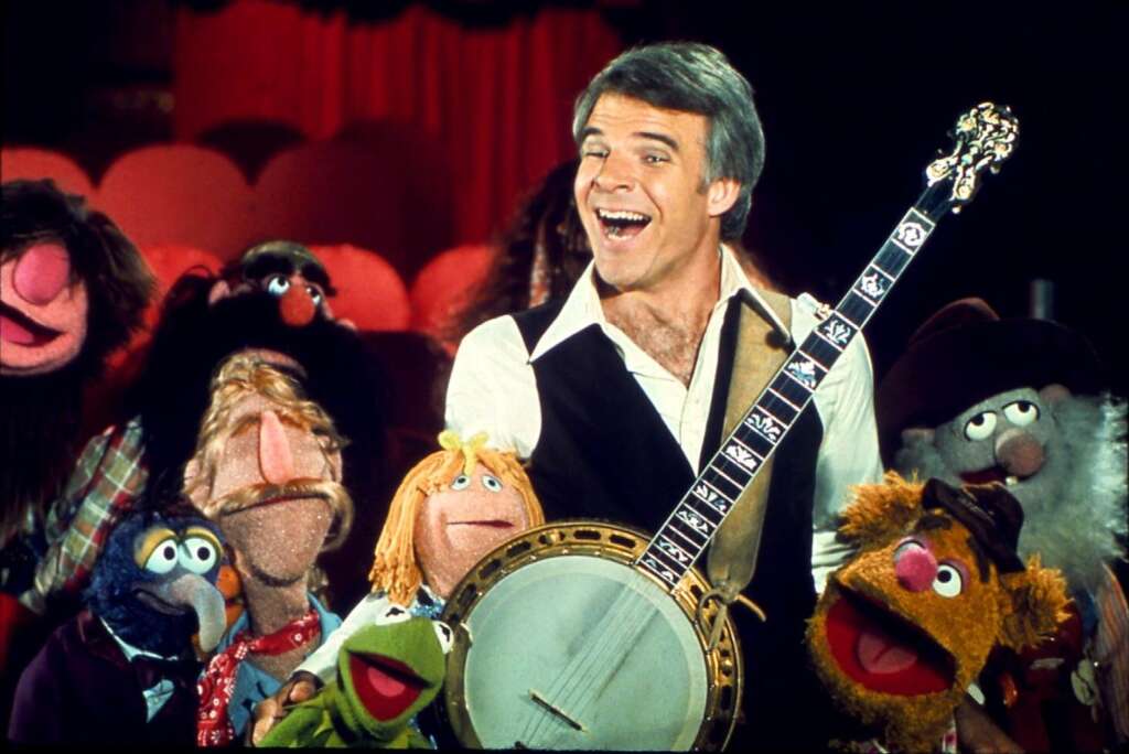 A smiling man holding a banjo stands in the center of a group of colorful puppets from The Muppet Show. The background features red theater seats and stage curtains. The puppets include Kermit the Frog, Miss Piggy, Fozzie Bear, and others.