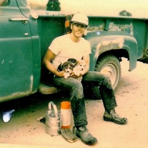 A man wearing a white t-shirt and cap is sitting on the ground next to an old green pickup truck. He is holding three small puppies in his lap and smiling. There's a thermos and a lunchbox on the ground beside him.