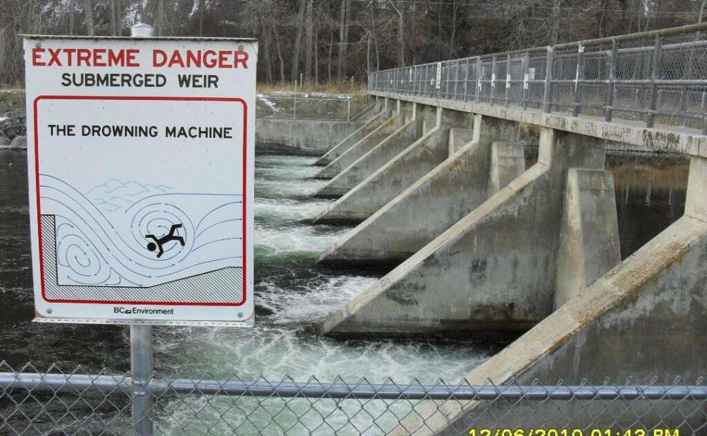 Warning sign next to a waterway with a concrete barrier structure and fast-flowing water. The sign reads "Extreme Danger: Submerged Weir - The Drowning Machine" with a graphic showing a person being pulled underwater. A chain-link fence runs along the front.